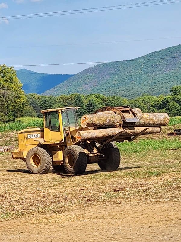 A yellow John Deere front loader lifts large logs on a dirt field with green hills and mountains in the background.