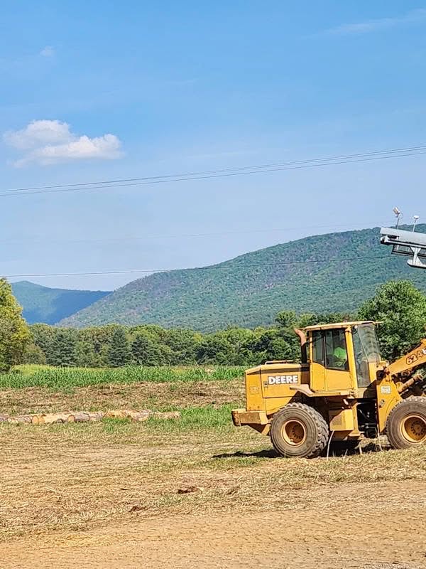 A yellow Deere front loader is parked on a cleared dirt field with green hills and a blue sky in the background.