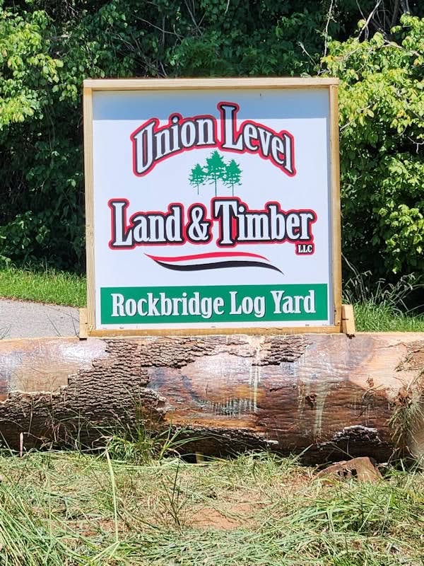 A sign reads "Union Level Land & Timber LLC, Rockbridge Log Yard," placed on a large log with greenery in the background.